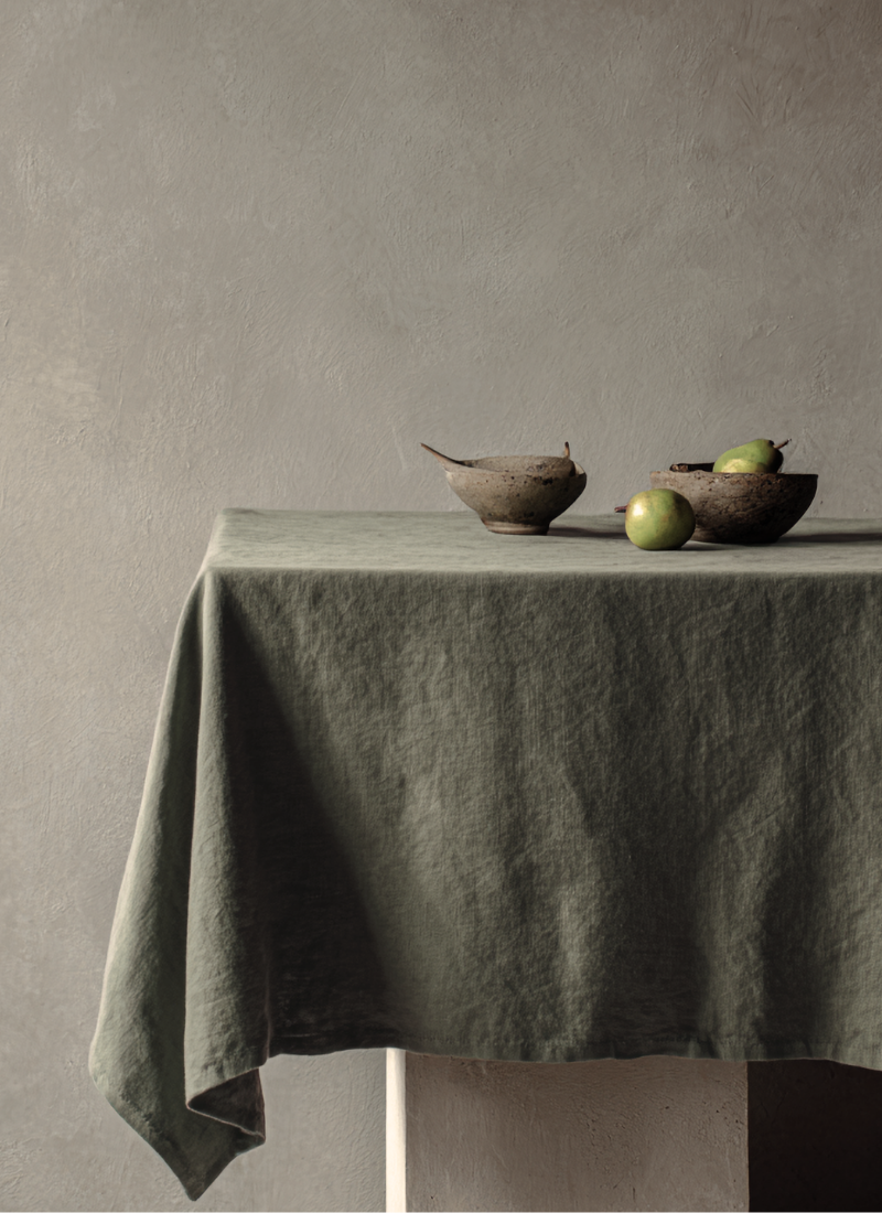 A table draped in a softly textured sage green linen tablecloth holding two rustic bowls and green pears, set against a textured grey wall.