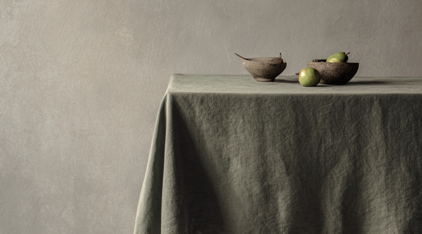 A table draped in a softly textured sage green linen tablecloth holding two rustic bowls and green pears, set against a textured grey wall.