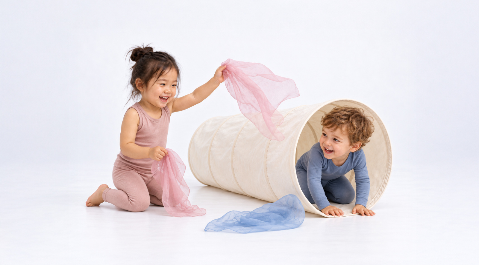 Two joyful toddlers play with colorful sheer scarves around a cream-colored fabric tunnel against a seamless white background. A little girl in a pink outfit waves a pink scarf while a smiling boy in blue crawls out of the tunnel.
