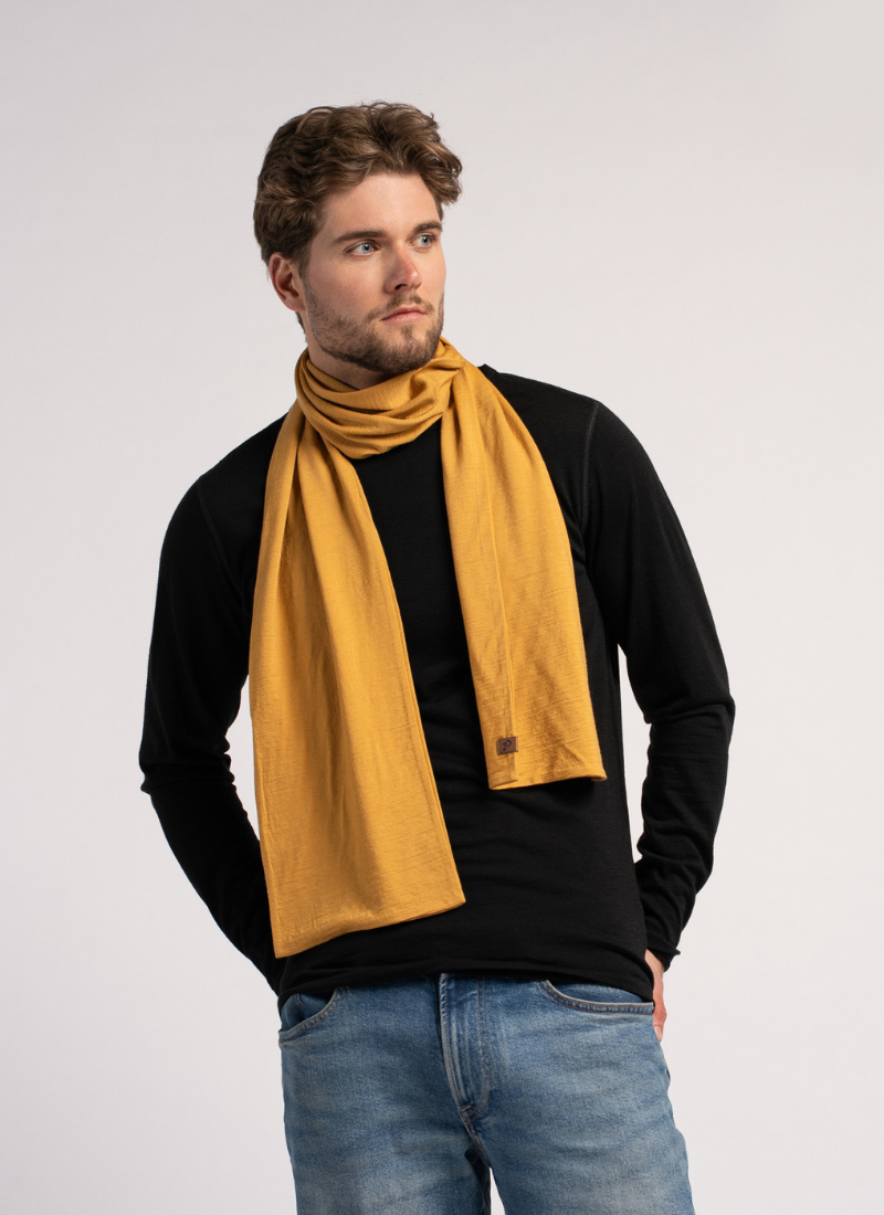 A man with a short beard stands against a light grey studio background wearing a mustard yellow knit scarf. He pairs the bright scarf with a black long-sleeved top and blue jeans while resting his hands in his pockets.