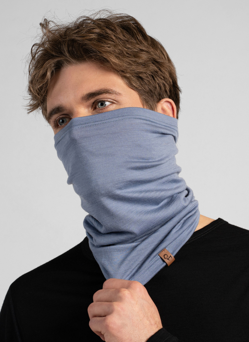 A man with wavy brown hair models a light blue knit neck gaiter pulled up to cover his nose and mouth. He wears a black long-sleeved shirt against a seamless light grey background, lightly holding the bottom edge of the gaiter.