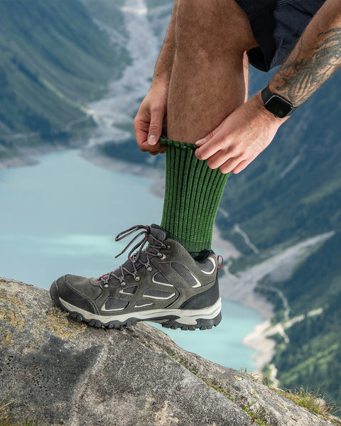 Man adjusting green ribbed Merino hiking socks while wearing hiking boots, scenic alpine lake view in the background.