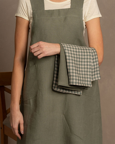 A woman holding two linen dish towels, one in stone green and the other in green check color, layered on top of one another in her hands. 