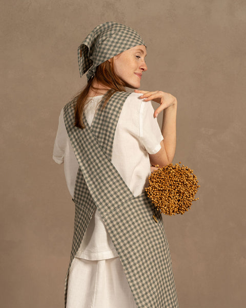 A person with fair skin and reddish-brown hair, possibly a young woman, smiles while looking over their shoulder, holding dried golden flowers. They wear a green checkered linen apron and headscarf.