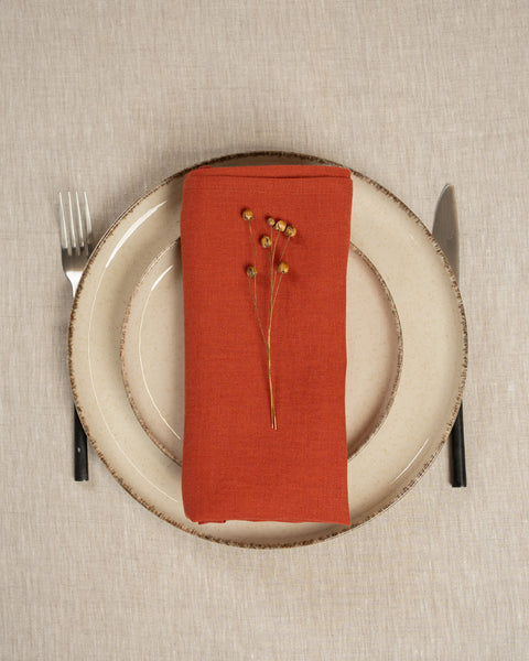 The menique linen napkin in cinnamon red color, put on top of two stacked plates . There are cutlery items by the sides of the plates. The background is a natural color linen tablecloth.