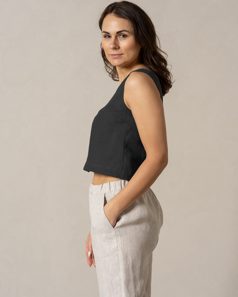 A woman with dark hair wearing 100% linen pants in the color natural and linen crop top in the color pure black. Her one hand is in the pocket. The background of the photo is in natural, nude colors.