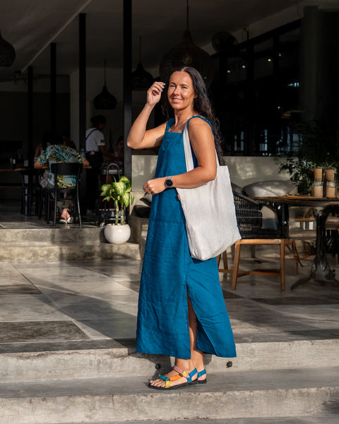 A smiling woman with long, dark hair is captured in a full outdoor shot, wearing a cobalt blue linen sundress with a square neckline, thick straps, and a side slit revealing colorful sandals. She has a menique linen tote bag slung over her shoulder and is touching her hair with one hand. The background features a building with open doorways showing interior tables, chairs, and a few people seated. Stone steps lead up to the building&#39;s entrance, and potted plants add touches of greenery.