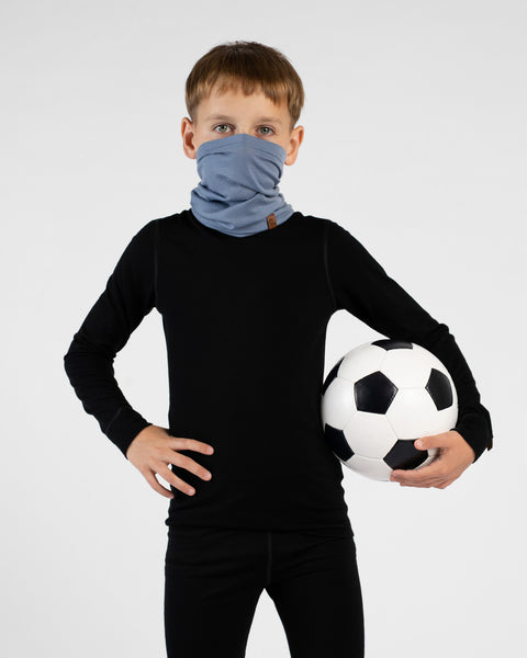 A young boy stands holding a black and white soccer ball under one arm with his other hand on his hip against a white background. He is wearing a black long-sleeved base layer top, black leggings, and a light blue neck gaiter pulled up over his nose and mouth.