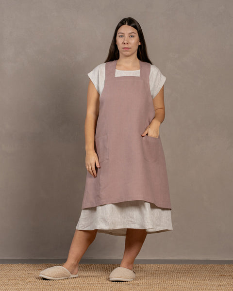 A woman wearing a faded rose color linen apron over a natural color menique linen dress, standing against a textured background.