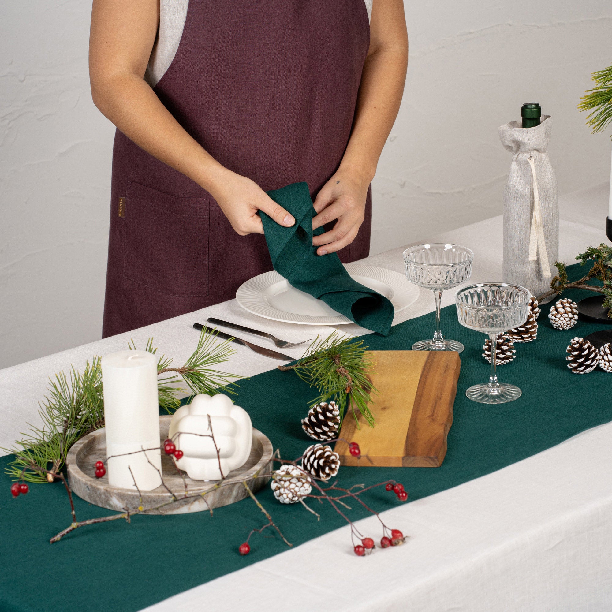 The menique linen tablecloth in dark green color, decorating a Christmas table. there are natural elements, such as pine cones and pine branches on the table top. There is also a wine bottle in a storage bag and candles.