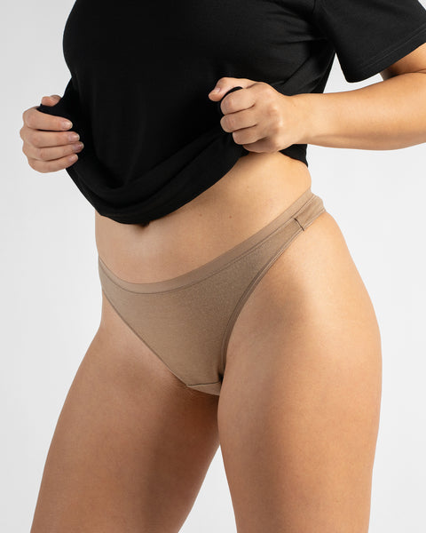 Three-quarter angle showing a woman holding up the hem of a black t-shirt to display seamless beige high-leg underwear bottoms.