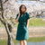 A woman with brown hair, wearing a dark green, short-sleeved wrap dress, stands outdoors under a tree with white blossoms.