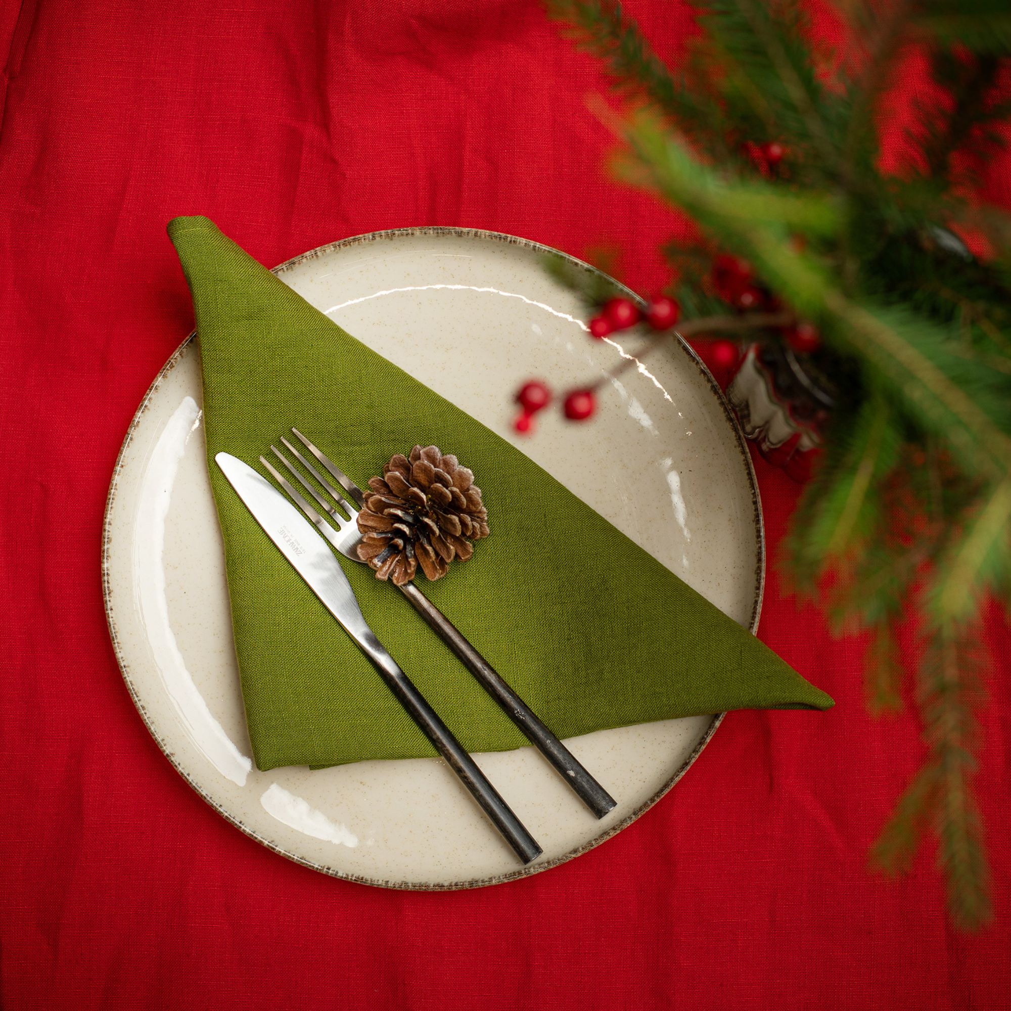 A decorated Christmas dinner table with a red table runner and green linen napkins folded on cream-colored plates. Pinecones, fir branches, red berries, and warm string lights create a festive centerpiece, with crystal glasses and red candles adding to the holiday ambiance. A Christmas tree glows in the background.