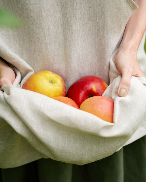 A close-up of hands gathering apples in the folds of a natural linen apron.