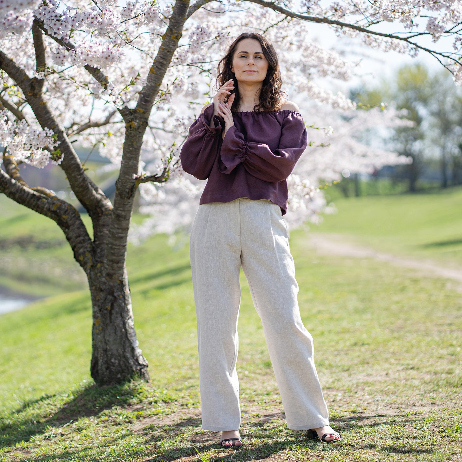 A woman with light skin and dark hair is shown from the waist up, smiling directly at the viewer. She is wearing a shadow purple, scoop-neck top. Her left hand, adorned with a ring on her ring finger, is casually raised to her chest, with her thumb touching her décolletage. 