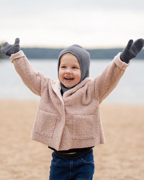 A young child with light skin and reddish-blonde hair is standing on a sandy beach, joyfully raising both arms with open hands. They are wearing a dark grey menique balaclava-style hat that covers their ears and neck, dark grey mittens, a light pink or peach-colored textured jacket, and dark blue jeans. Their mouth is open in a wide smile, showing teeth, and their eyes are sparkling. In the background, there is a body of water under a light sky.