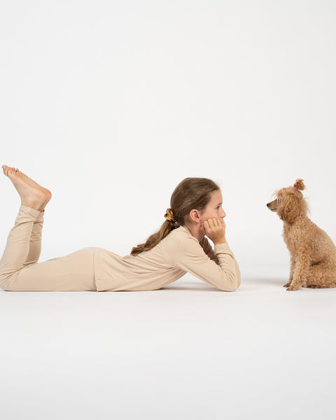 Girl lying on the floor in a beige bamboo long-sleeve top and bottoms, facing a small brown poodle sitting in front of her.