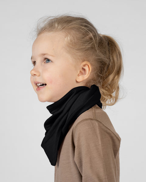 A studio portrait of a young blonde girl looking upwards and smiling slightly. She is seen in profile from the shoulders up. She is wearing a solid black bandana tied around her neck, over a light brown long-sleeved shirt. The background is a plain light grey.