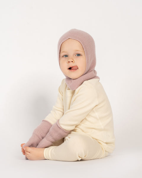 A young child with blue eyes, is sitting on a white surface, sticking out their tongue and looking directly at the viewer. They are wearing a pink balaclava, matching pink mittens, and a light-colored long-sleeved shirt and pants.