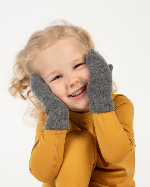 A young child with curly blonde hair and a bright smile wears a spicy yellow long-sleeved shirt and dark gray double mittens, with their hands held up to their cheeks. The background is white.