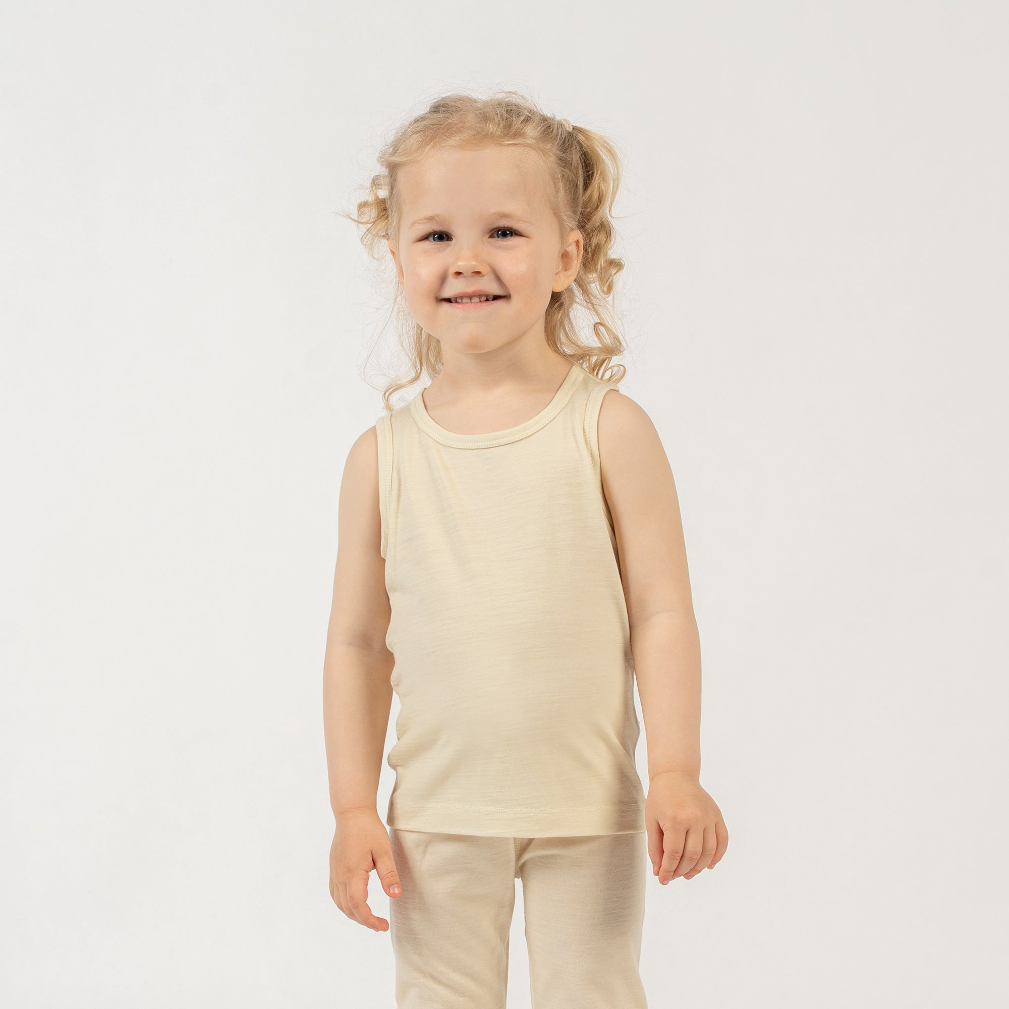 A young child with curly brown hair stands in front of a dark, textured wall with a modern fireplace opening. The child is wearing a natural sleeveless top and matching pants. They are looking directly at the camera with a slight smile, with one hand on their hip.