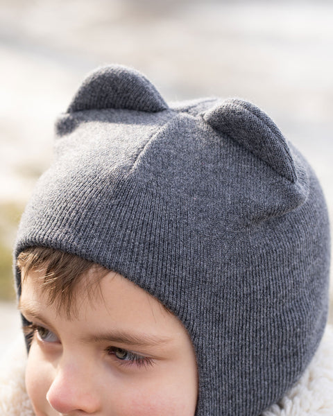 A close-up of a child&#39;s head, partially obscured, wearing a dark gray menique knit balaclava or hat with two ear-like protrusions on top. The child&#39;s right eye and part of their face are visible, and they appear to be looking slightly to their right. A textured, light-colored collar or hood of a jacket is visible around their neck, and the background is blurred with light tones, suggesting an outdoor setting.