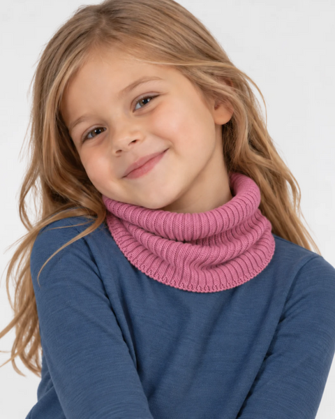 Young girl with long blonde hair tilting her head and smiling at the camera, wearing a pink ribbed knit neck gaiter and a blue long-sleeve top against a white background.
