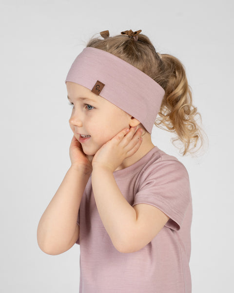 A studio portrait of a smiling young child in a three-quarter profile facing left, against a plain light grey background. She is wearing a light dusty pink short-sleeved t-shirt and a matching wide dusty pink headband with a small brown logo tag, gently holding her hands to her cheeks.