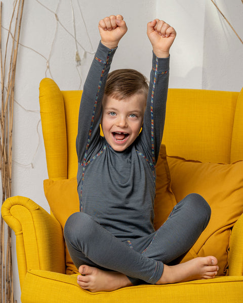 A boy sitting in a yellow armchair, wearing the menique Kids&#39; 160 Merino Pants in Perfect Grey color. He has his hands raised above his head, being cheerful.