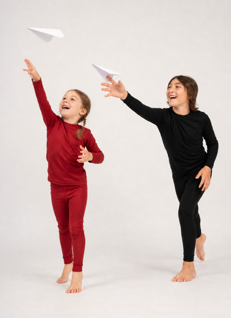 Two joyful children barefoot against a white background, one in a red base layer set and the other in black, reaching upward toward flying paper airplanes.