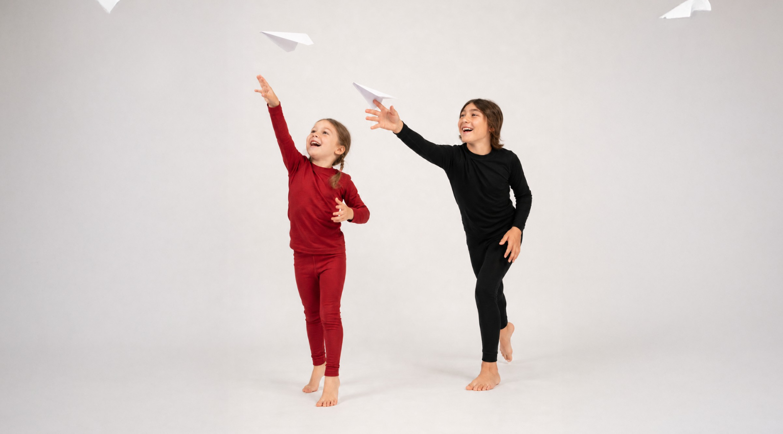 Two joyful children barefoot against a white background, one in a red base layer set and the other in black, reaching upward toward flying paper airplanes.
