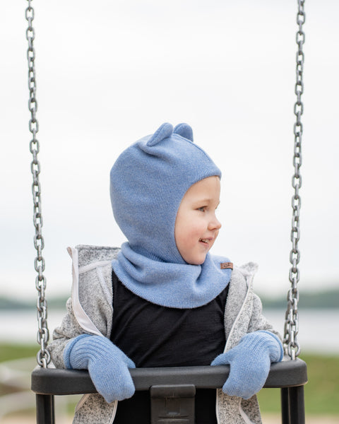 A young child with blonde hair, is sitting in a playground swing and looking to the right with a slight smile. They are wearing a light blue balaclava with small ear-like features, light blue mittens, a grey speckled zip-up hoodie, and a black shirt. The background is a blurred outdoor scene with water and land.