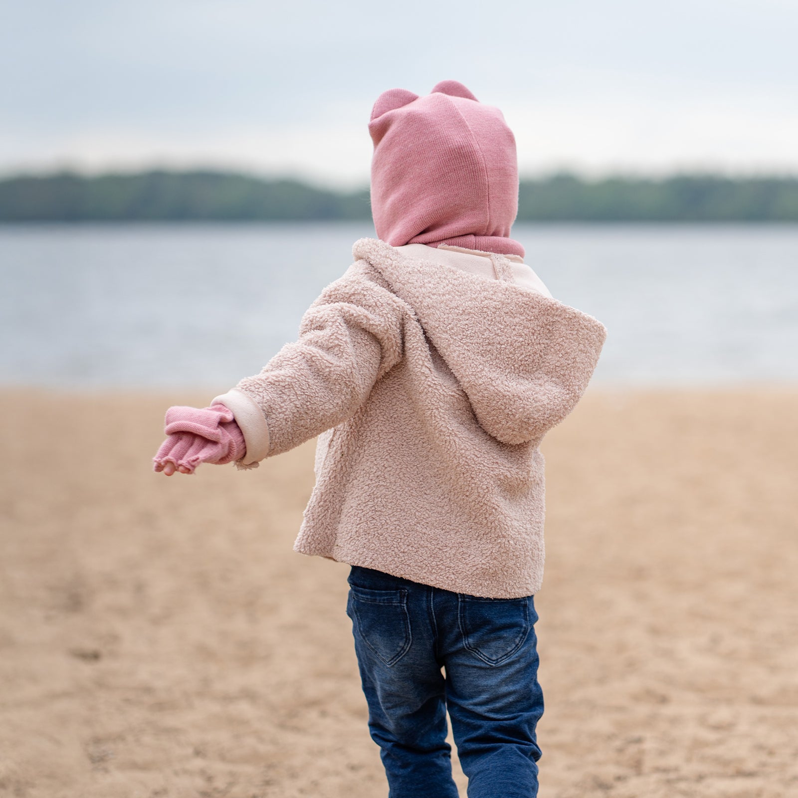 A joyful child wearing a pink balaclava with ears, a pink scarf, and a beige coat stands on a beach, hands open in excitement.