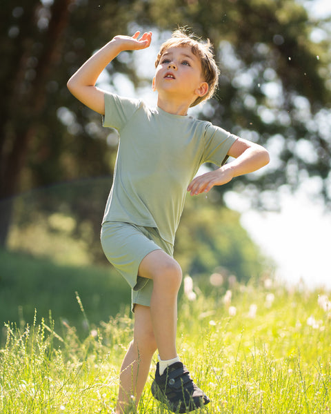 Young boy in a sunny meadow wearing a light green bamboo T-shirt and matching shorts. The lightweight kids' outfit is showcased in a natural outdoor setting, highlighting its soft, breathable comfort for everyday adventures.