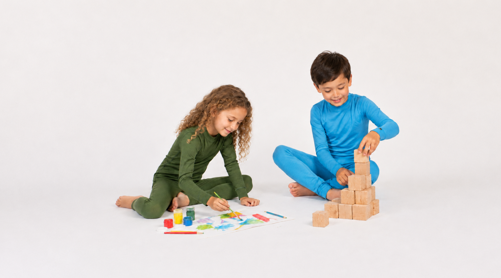 Two barefoot children sitting on a white floor playing. A girl on the left wearing an olive green long-sleeved base layer set paints a colorful picture, while a boy on the right wearing a blue long-sleeved base layer set builds a pyramid out of wooden blocks.