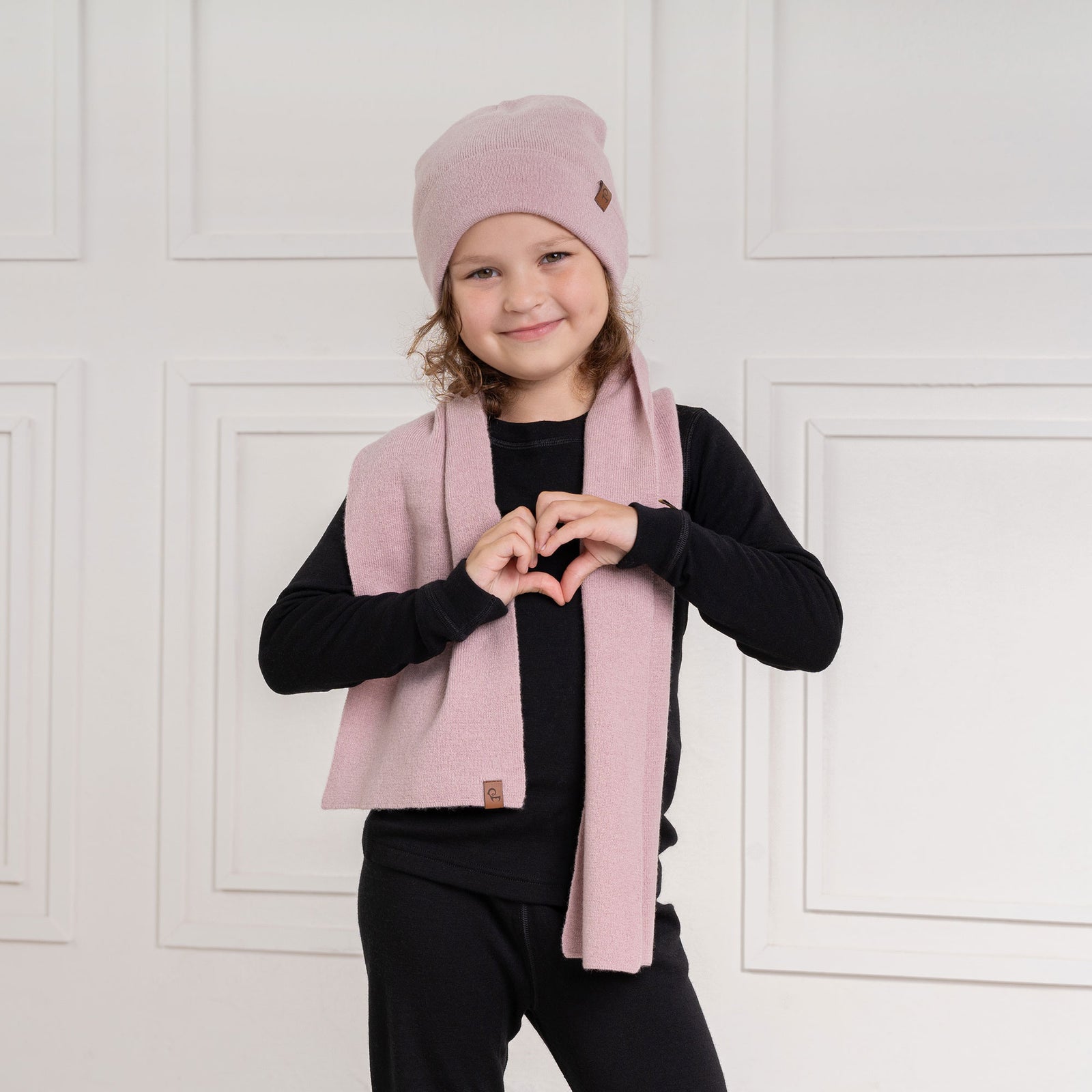 A young child with curly brown hair and a slight smile looks directly at the viewer. They are wearing a black long-sleeved shirt and a light pink scarf with a small brown label, draped around their neck. The background is a white paneled wall.