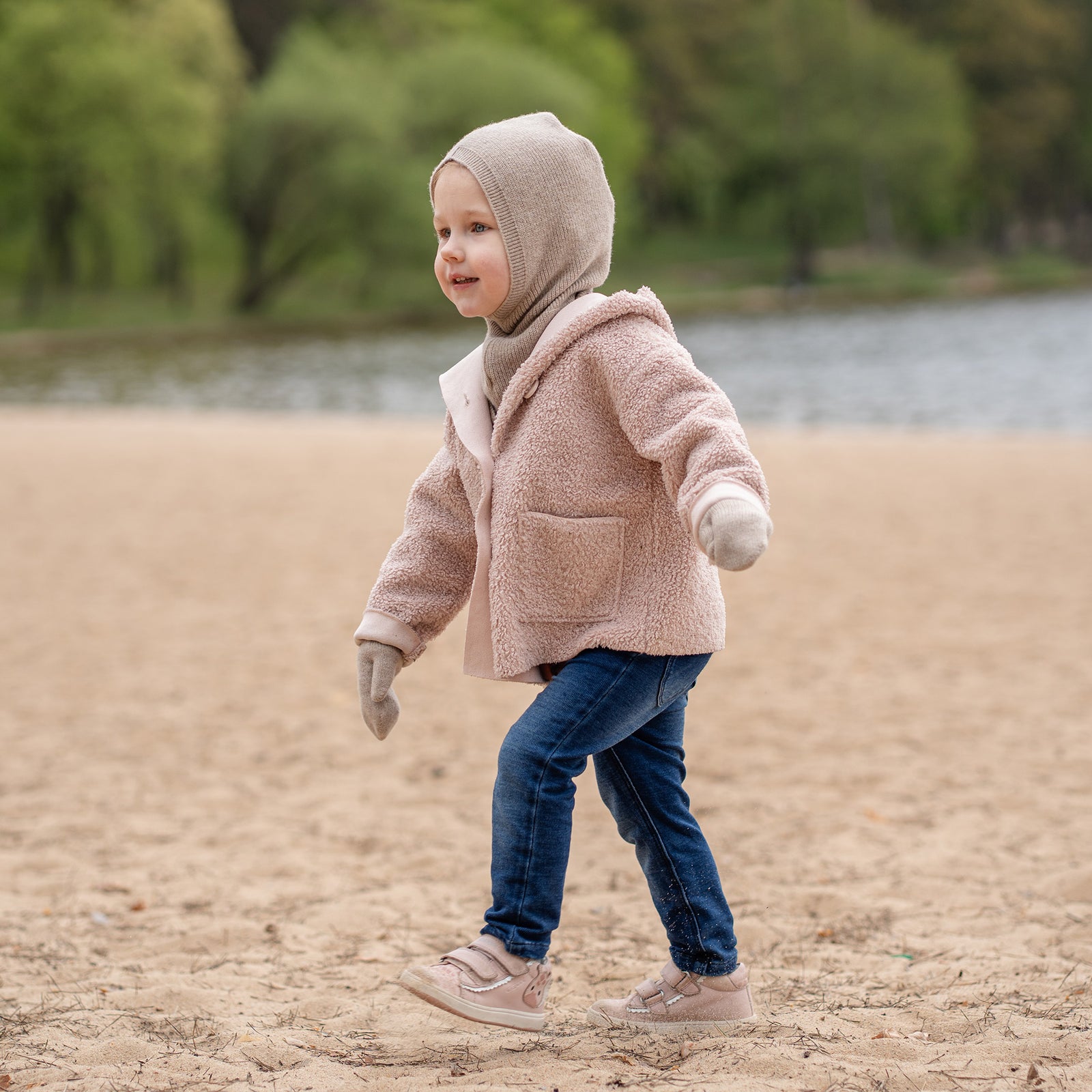 A happy young child with blonde hair and blue eyes, stands on a sandy beach. They are wearing a beige balaclava, a matching beige scarf, a light pink sherpa-style coat, beige mittens, and blue jeans, looking directly at the camera with a blurred body of water in the background.