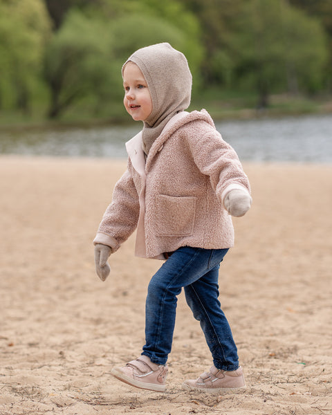 A young child is walking on a sandy beach, looking to the right with a slight smile. They are dressed in a beige balaclava, a matching beige scarf, a light pink sherpa-style coat, beige mittens, blue jeans, and pink sneakers. A blurred body of water and green trees are in the background.