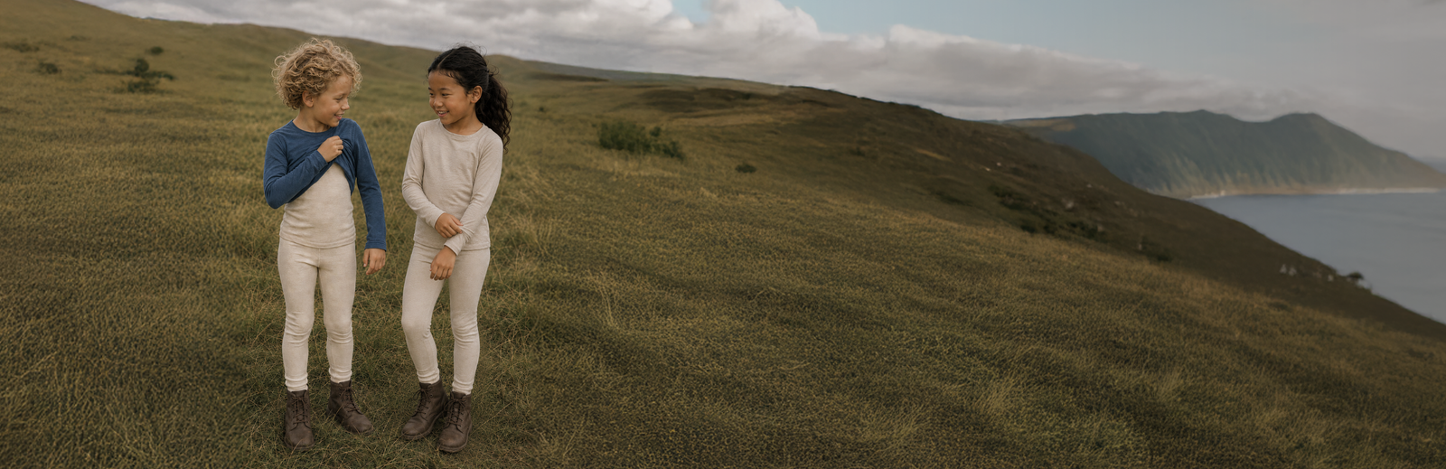 Two young children in long-sleeved base layers standing together in a grassy field under a partly cloudy sky.