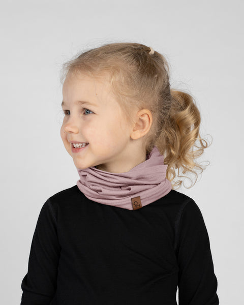 A studio portrait of a smiling young blonde girl looking off to the left side of the frame. Her blonde hair is pulled back into a ponytail. She is wearing a dusty pink neck gaiter gathered around her neck, which has a small brown leather logo patch near the bottom edge. She is wearing a solid black long-sleeved shirt against a plain light grey background.