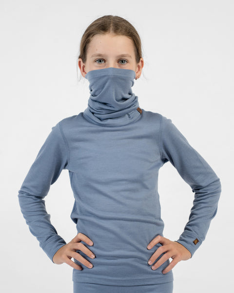 A young girl with her hands on her hips, wearing a dusty blue long-sleeve shirt and a matching dusty blue merino long neck gaiter pulled up to cover her nose and mouth.