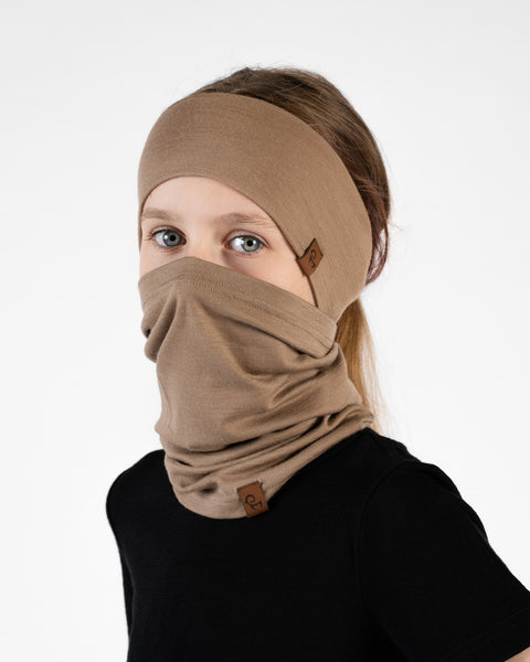 A close-up portrait of a young girl looking directly at the camera in a three-quarter profile against a white background. She is wearing a black short-sleeved t-shirt, a beige headband, and a matching beige neck gaiter pulled up over her nose and mouth.