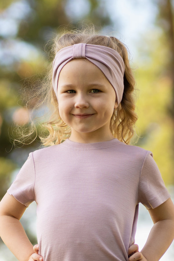 A young, fair-skinned girl with wavy blonde hair smiles gently at the camera while standing outdoors with her hands resting on her hips. She is wearing a light dusty pink short-sleeved t-shirt and a matching dusty pink fabric headband with a gathered, ribbon-like center detail. The background is a softly blurred outdoor setting with trees and dappled sunlight.
