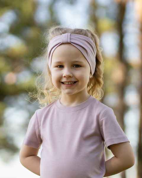 Young girl smiling outdoors wearing a dusty pink Merino wool T-shirt and a matching Merino wool headband. The soft, breathable accessories and clothing are showcased in a sunny natural setting, offering comfortable everyday wear for active ki