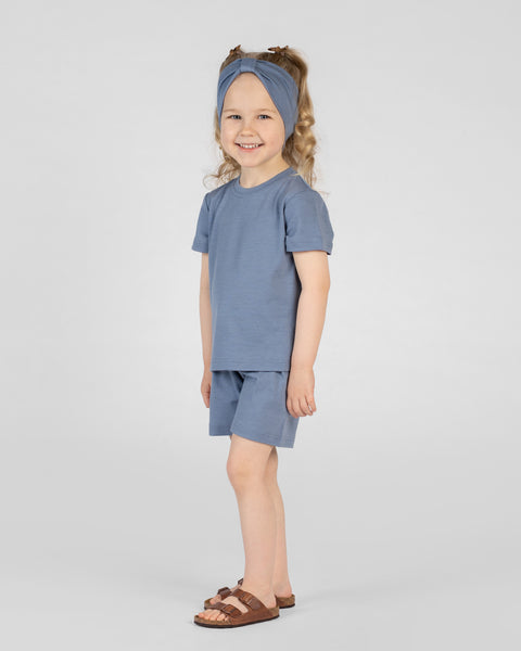 A young, smiling girl stands slightly in profile while looking towards the camera against a plain light gray background. She is wearing a matching dusty blue outfit consisting of a short-sleeved t-shirt, shorts, and a wide fabric headband with a center knot detail, paired with brown double-strap sandals.