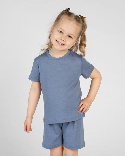 A young child poses playfully with one hand on their hip and a bright smile, wearing a dusty blue short-sleeve t-shirt and matching shorts. They have blonde hair styled in half-up pigtails with small brown clips, set against a plain light background.