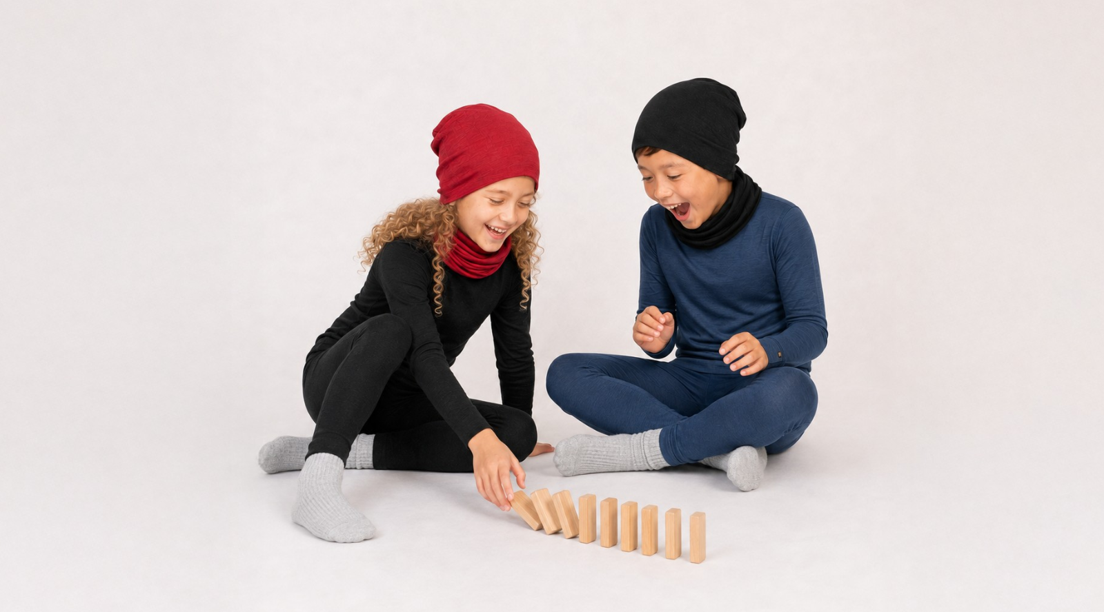 Two children smiling and sitting on a white floor playing with wooden dominoes. The girl on the left is dressed in a black long-sleeved base layer set, a dark red beanie, a matching red neck gaiter, and grey ribbed socks. The boy on the right wears a navy blue long-sleeved base layer set, a black beanie, a matching black neck gaiter, and grey ribbed socks.