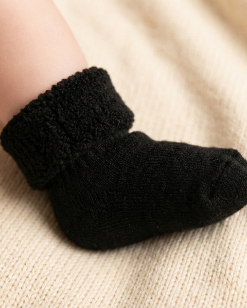 Close-up side view of a baby wearing black sherpa-lined socks with a soft knitted foot and fluffy folded cuff, photographed on a neutral fabric surface to highlight warmth and comfort.