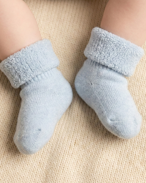 Close-up of a baby’s feet wearing light blue sherpa-lined socks, featuring a soft knit exterior and plush folded cuffs, shown on a neutral textured blanket for a cozy, warm look.