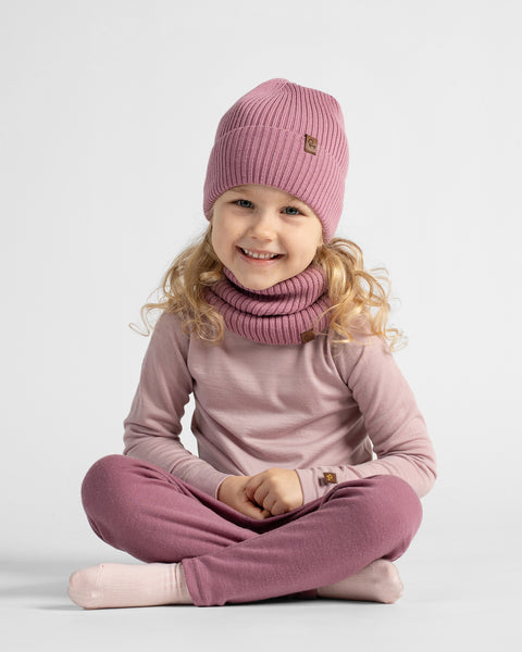A full-length view of a young girl sitting cross-legged on the floor, wearing a mauve loungewear set paired with a matching pink knit beanie and snood.